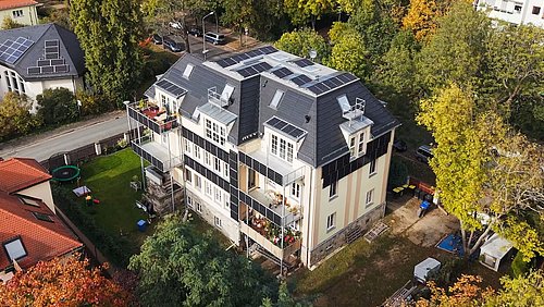 Foto, mehrstöckige Stadtvilla mit mehreren Balkonen und zahlreichen Solarpaneelen auf dem Dach umgeben von Bäumen und Nachbarhäusern.