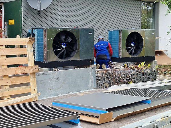 Foto, Zwei große Außeneinheiten mit Wärmepumpen Ventilatoren vor einer Gebäudewand, daneben eine Person in Arbeitskleidung, im Vordergrund Paletten und Metallgitter