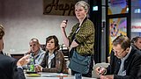 Foto, Frau mit grauem Haar und gemustertem Cardigan steht mit Mikrofon in der Hand vor sitzenden Personen an einem Tisch mit Getränken und Notizmaterial.
