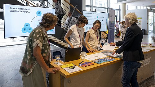 Foto, drei Frauen hinter einem Messetisch mit Informationsmaterial und Laptop sprechen mit einer Besucherin in einem hellen Raum mit Treppe und Monitoren im Hintergrund.