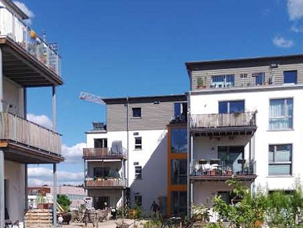 Foto, Mehrgeschossige Wohnhäuser mit Balkonen und großen Fenstern stehen unter blauem Himmel. Im Vordergrund sind Pflanzen und Sitzgelegenheiten zu sehen, die auf einen gemeinschaftlich genutzten Außenbereich hindeuten. Die Gebäude wirken modern und hell, mit Holz- und Metallakzenten an den Balkonen.