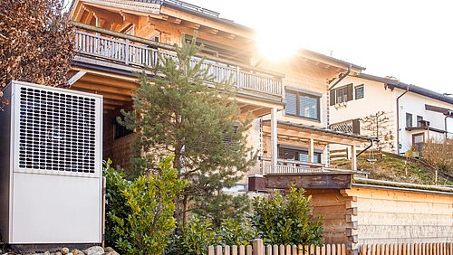 Foto, Holzhaus mit Balkon und Garten, im Vordergrund Wärmepumpe neben einem kleinen Zaun und Büschen