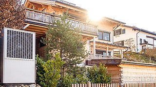 Foto, Holzhaus mit Balkon und Garten, im Vordergrund Wärmepumpe neben einem kleinen Zaun und Büschen