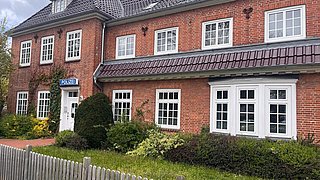 Zweistöckiges rotes Backsteingebäude mit mehreren weißen Fenstern, einem Schild mit der Aufschrift 'POLIZEI' und einem kleinen Garten mit Sträuchern davor.