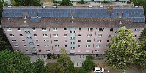 Foto, Mehrstöckiges Wohngebäude mit Solarpaneelen auf dem Dach, umgeben von Bäumen und Parkplätzen mit geparkten Autos