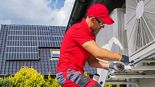 Foto, im Vordergrund ein Mann auf einer Leiter, der Wärmepumpen prüft, im Hintergrund ein Haus mit einer Solaranlage auf dem Dach.