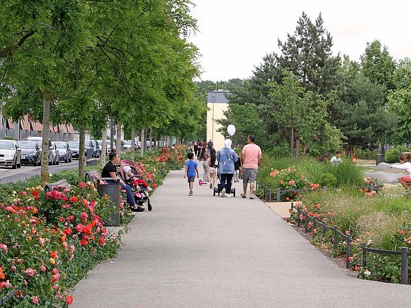 Foto, Breiter Fußweg mit blühenden Rosenbeeten und mehreren Personen, darunter eine Familie mit Kinderwagen, in einem Park oder einer Grünanlage