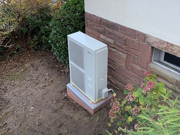 Foto, Außenwand eines Hauses mit einer weißen Wärmepumpe auf einem Betonfundament neben einem Fenster