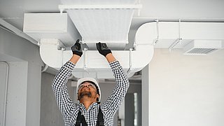 Foto, Ein Arbeiter im karierten Hemd und mit Handschuhen arbeitet an einer Klimaanlage an der Decke eines modernen, unfertigen Raums.