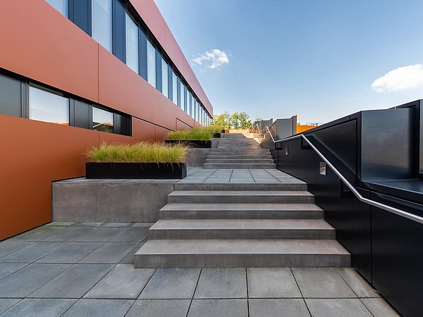 Foto, Breite Außentreppe mit grauen Stufen und schwarzem Geländer neben einem modernen Gebäude mit orangen Fassadenelementen