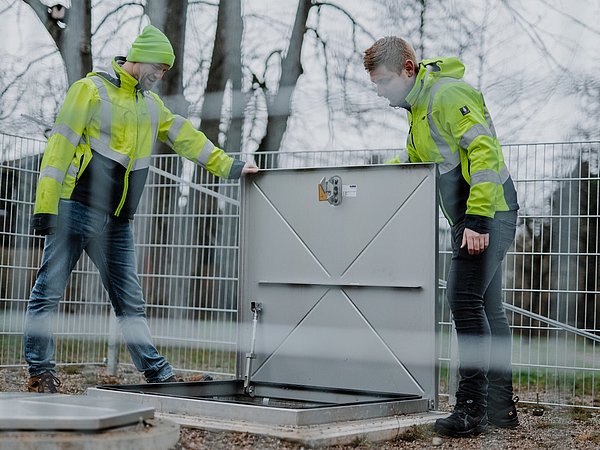 Zwei Personen in gelb-grünen Warnjacken öffnen eine Metallklappe auf einem umzäunten Gelände mit Bäumen im Hintergrund.