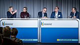Foto, fünf Personen sitzen an zwei nebeneinanderstehenden Tischen mit der Aufschrift "Gebäudeforum Klimaneutral", vor ihnen stehen Gläser mit Wasser, im Vordergrund Publikum.