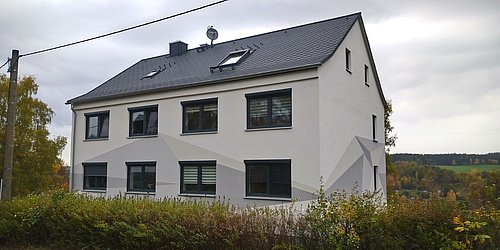 Foto, Zweistöckiges Wohnhaus mit grauem Satteldach und mehreren Fenstern, umgeben von Herbstbüschen und Bäumen