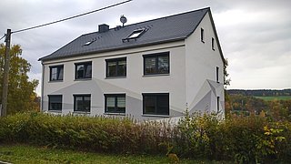 Foto, Zweistöckiges Wohnhaus mit grauem Satteldach und mehreren Fenstern, umgeben von Herbstbüschen und Bäumen