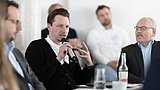 Foto, Person hält Mikrofon und spricht in einer Gesprächsrunde mit mehreren Personen
