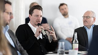 Foto, Person hält Mikrofon und spricht in einer Gesprächsrunde mit mehreren Personen