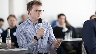 Foto, Mann mit Brille und gestreiftem Hemd hält Mikrofon und spricht in einem Raum mit mehreren Personen im Hintergrund.