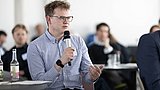 Foto, Mann mit Brille und gestreiftem Hemd hält Mikrofon und spricht in einem Raum mit mehreren Personen im Hintergrund.