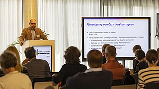 Foto, ein Redner hält einen Vortrag auf einer Konferenz zum Thema Klimaneutralität mit einer informativen Folie über Nachbarschaftskonzepte und Herausforderungen.