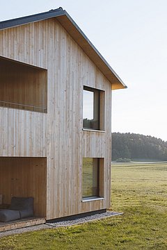 Foto, zweistöckiges Holzhaus mit Balkon, großen Fenstern und Sitzbereich auf der Terrasse, umgeben von Wiese und Wald im Hintergrund.