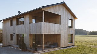 Foto, zweistöckiges Holzhaus mit Balkon, großen Fenstern und Sitzbereich auf der Terrasse, umgeben von Wiese und Wald im Hintergrund.
