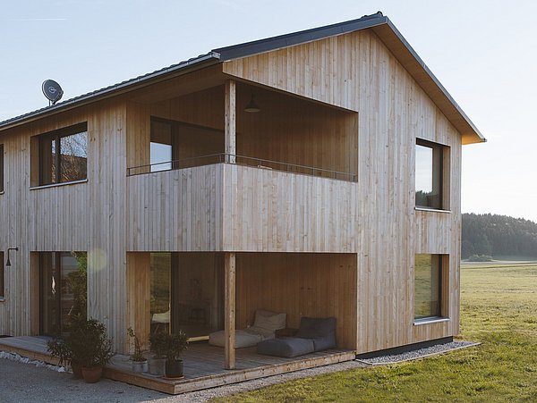Zweistöckiges Holzhaus mit Balkon, großen Fenstern und Sitzbereich auf der Terrasse, umgeben von Wiese und Wald im Hintergrund.
