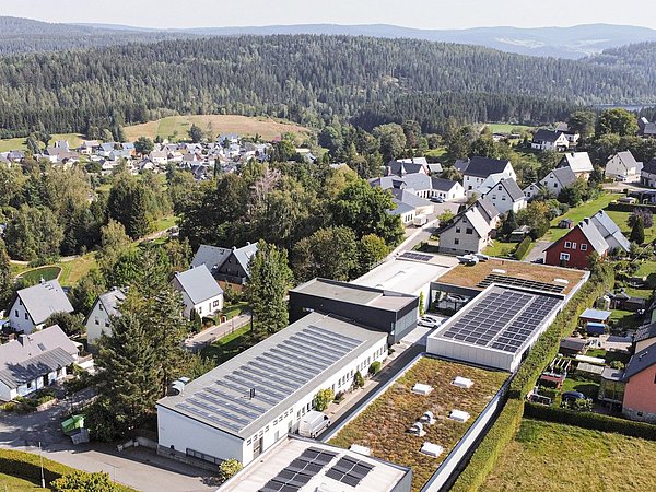 Foto, Luftaufnahme mit der Produktionsstätte, Wohnhäusern und einem großen Gebäude mit begrüntem Dach und Solarpaneelen.