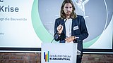 Foto, Person mit langen Haaren und dunklem Blazer spricht am Rednerpult mit Logo 'Gebäudeforum Klimaneutral' bei Veranstaltung