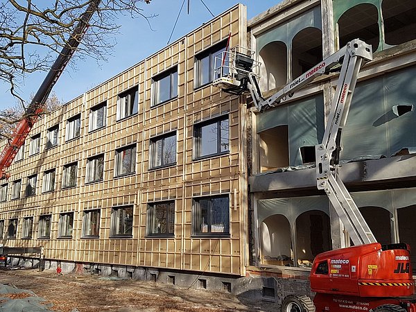 Foto, Baustelle des Verwaltungsgebäudes mit auf der Fassade angebrachter Dämmung und eine Hebebühne im Vordergrund.