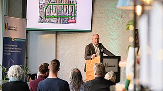 Foto, Mann steht an einem Rednerpult vor Publikum und präsentiert eine Karte auf einem Bildschirm an der Wand.