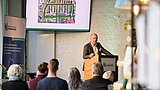 Foto, Mann steht an einem Rednerpult vor Publikum und präsentiert eine Karte auf einem Bildschirm an der Wand.