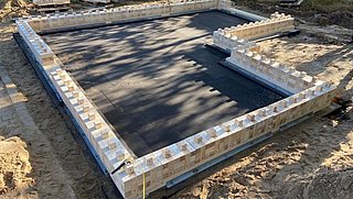 Foto, Baustelle mit einem schwarzen Fundamentsockel, umgeben von Holzblöcken auf einer sandigen Oberfläche, mit Werkzeugen und Materialien in der Nähe.