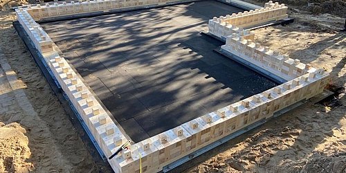 Foto, Baustelle mit einem schwarzen Fundamentsockel, umgeben von Holzblöcken auf einer sandigen Oberfläche, mit Werkzeugen und Materialien in der Nähe.