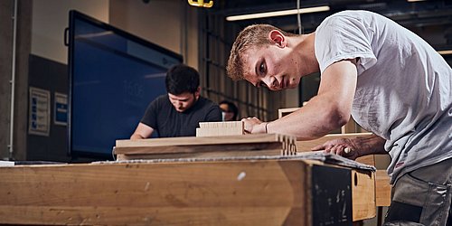 Foto, eine Person arbeitet gebeugt über einer Werkbank. Im Hintergrund befindet sich eine weitere Person. 