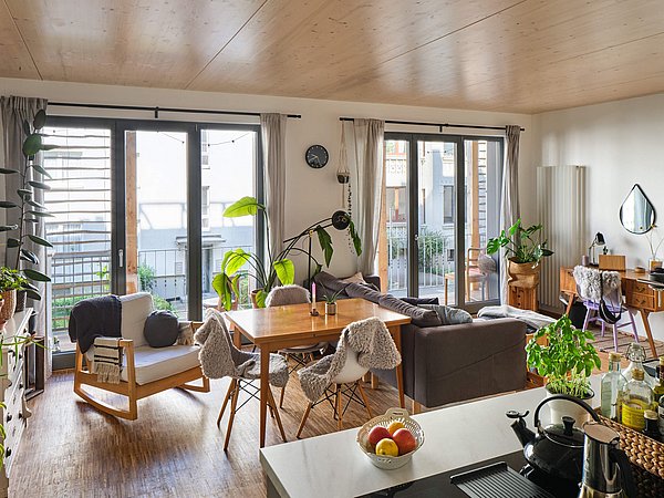 Foto, Heller Wohnraum mit Holzboden, Esstisch mit Stühlen, Pflanzen und großen Fenstern mit Blick auf Balkone