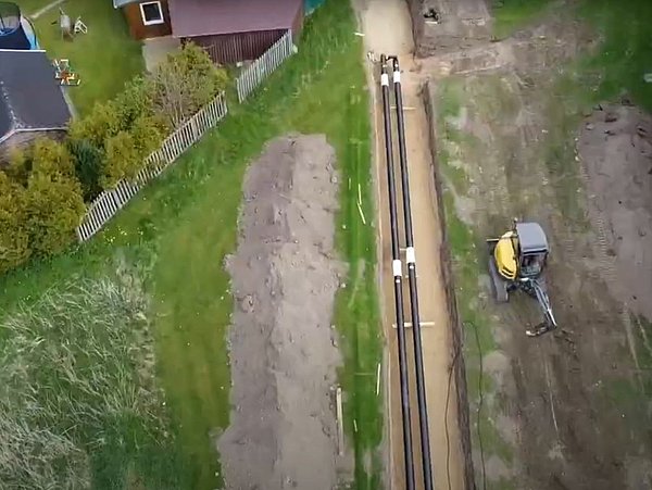 Foto, Luftaufnahme einer Baustelle mit einem kleinen Bagger, neu verlegten Rohren und umgebender Vegetation, darunter ein Trampolin im Hof.