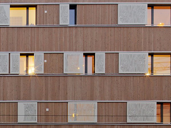 Foto, Gebäudefassade aus Holz mit Fenstern und Schiebeelementen vor einigen Fenstern.