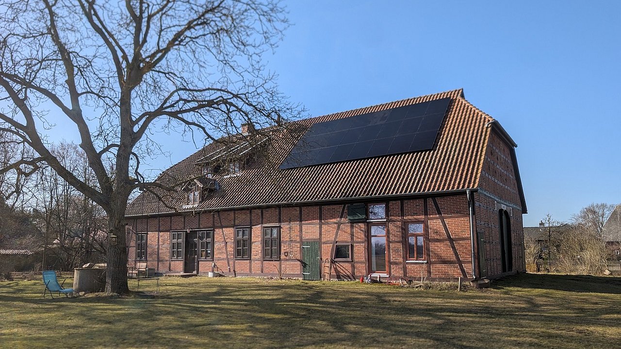 Foto, Großes Fachwerkhaus mit Ziegeldach und Solarpaneelen auf der Dachhälfte, umgeben von Rasen und Bäumen