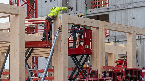 Foto, zwei Bauarbeiter mit Schutzhelmen und Warnkleidung montieren Holzelemente auf einer Baustelle mit Betongebäude im Hintergrund.