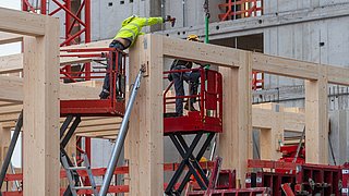 Foto, zwei Bauarbeiter mit Schutzhelmen und Warnkleidung montieren Holzelemente auf einer Baustelle mit Betongebäude im Hintergrund.