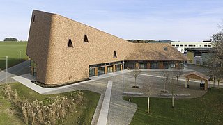 Foto, Besucherzentrum mit geschwungenem, strohgedecktem Dach und kleinen dreieckigen Fenstern, umgeben von Wegen und Bäumen