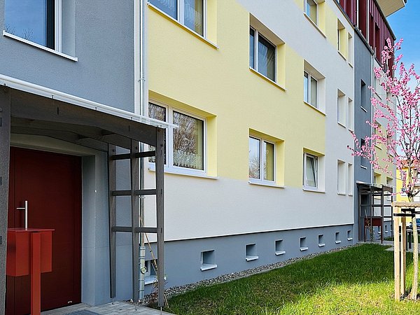 Eingang eines mehrstöckigen Wohnhauses mit roten Türen, gelb-grauer Fassade und blühendem Baum im Vorgarten.