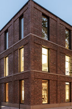 Foto, Ein modernes, dreistöckiges Gebäude aus dunkelrotem Backstein steht an einer gepflasterten Fläche. Die großen, rechteckigen Fenster sind gleichmäßig angeordnet und leuchten von innen. Links im Bild sind einige Straßenlaternen zu sehen, rechts wachsen mehrere hohe Bäume mit grünen Blättern.