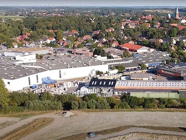 Foto, Luftaufnahme eines großen Industriekomplexes, umgeben von Bäumen und Wohngebäuden, mit Lagerhallen und angrenzenden Feldern.