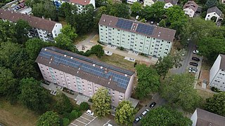 Foto, Mehrfamilienhäuser mit Solaranlagen auf den Dächern, umgeben von Bäumen und Parkplätzen in einem Wohngebiet
