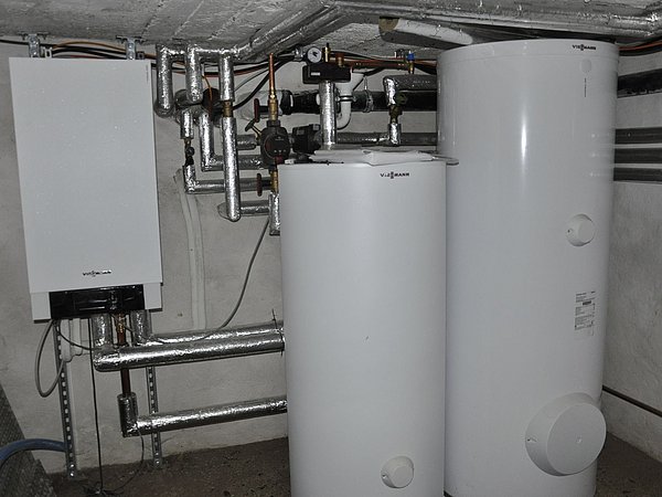 Foto, Heizungsanlage mit zwei weißen Warmwasserspeichern und vernetzten Rohrleitungen in einem Keller