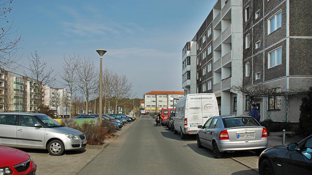 Foto, Blick in eine Wohnstraße mit geparkten Autos auf beiden Seiten und mehrstöckigen Wohngebäuden bei klarem Himmel