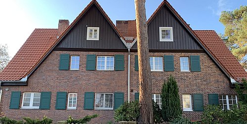 Foto, Zweistöckiges Doppelhaus mit roten Ziegeldächern, braunen Holzgiebeln und grünen Fensterläden, davor ein Zaun und ein Baum
