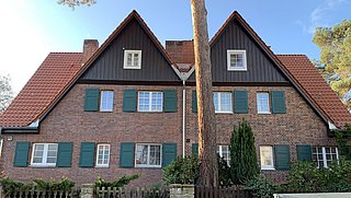 Foto, Zweistöckiges Doppelhaus mit roten Ziegeldächern, braunen Holzgiebeln und grünen Fensterläden, davor ein Zaun und ein Baum