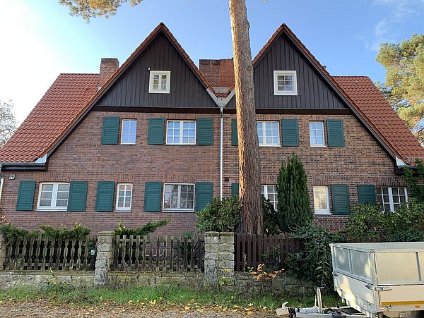 Foto, Zweistöckiges Doppelhaus mit roten Ziegeldächern, braunen Holzgiebeln und grünen Fensterläden, davor ein Zaun und ein Baum